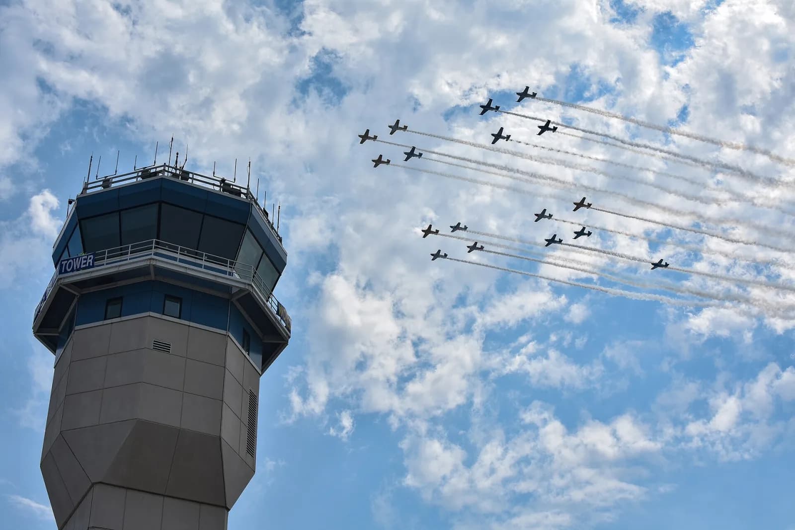 Formação de cerca de vinte aviões a hélice soltando fumaça branca ao lado da torre de controle do aeroporto, sob céu azul com nuvens
