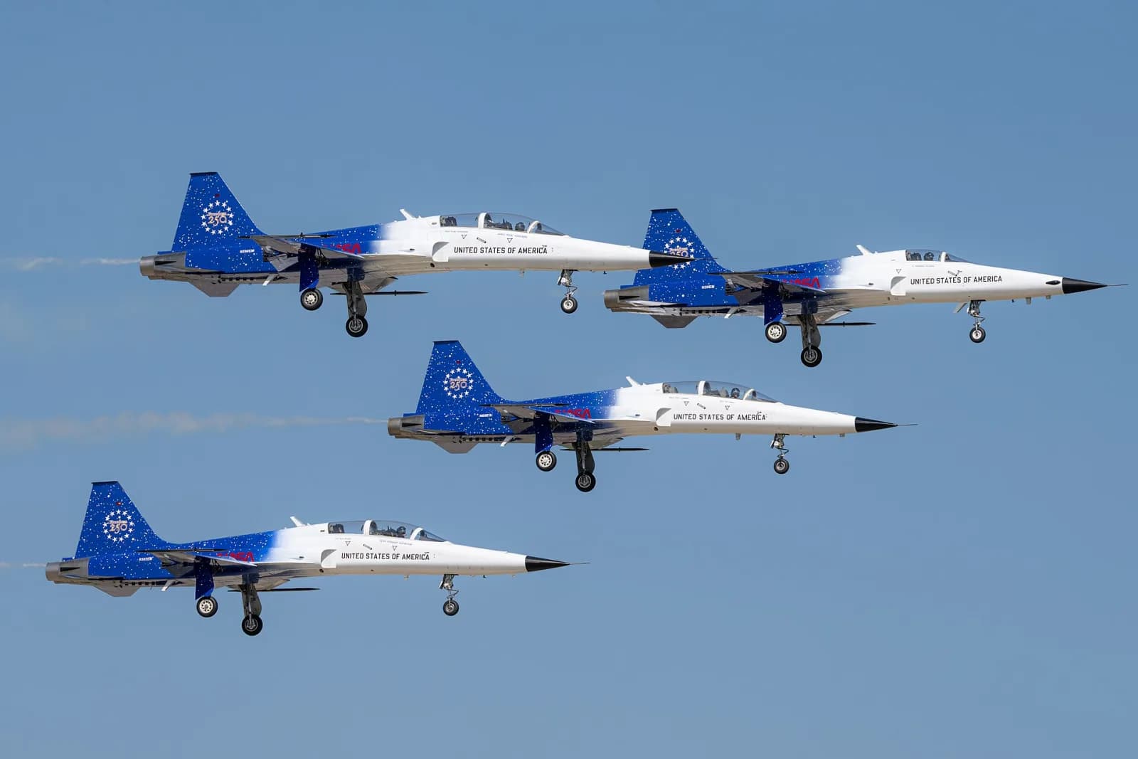 Quatro jatos brancos e azuis com estrelas, a inscrição United States of America e o emblema Freedom 250 na deriva, voando juntos com o trem de pouso baixado