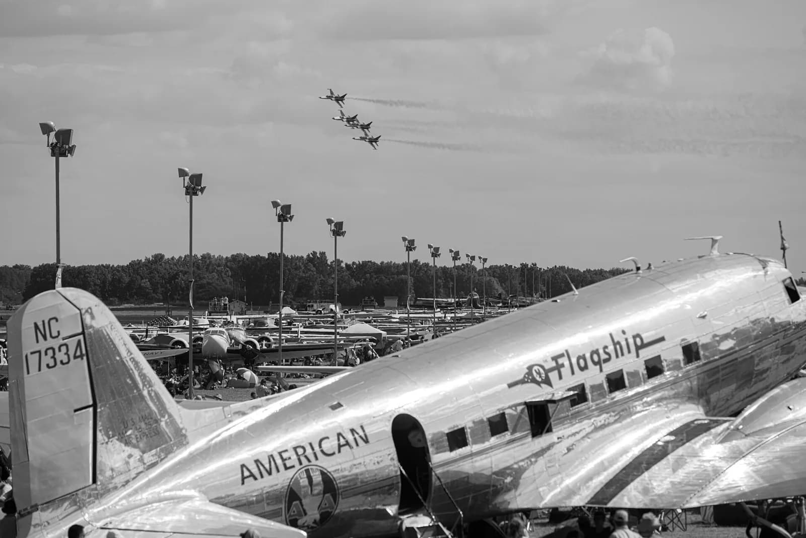 Foto em preto e branco de quatro jatos em formação no céu, acima de um pátio cheio de aviões antigos e de público, com um bimotor clássico de alumínio polido com a inscrição American Flagship em primeiro plano
