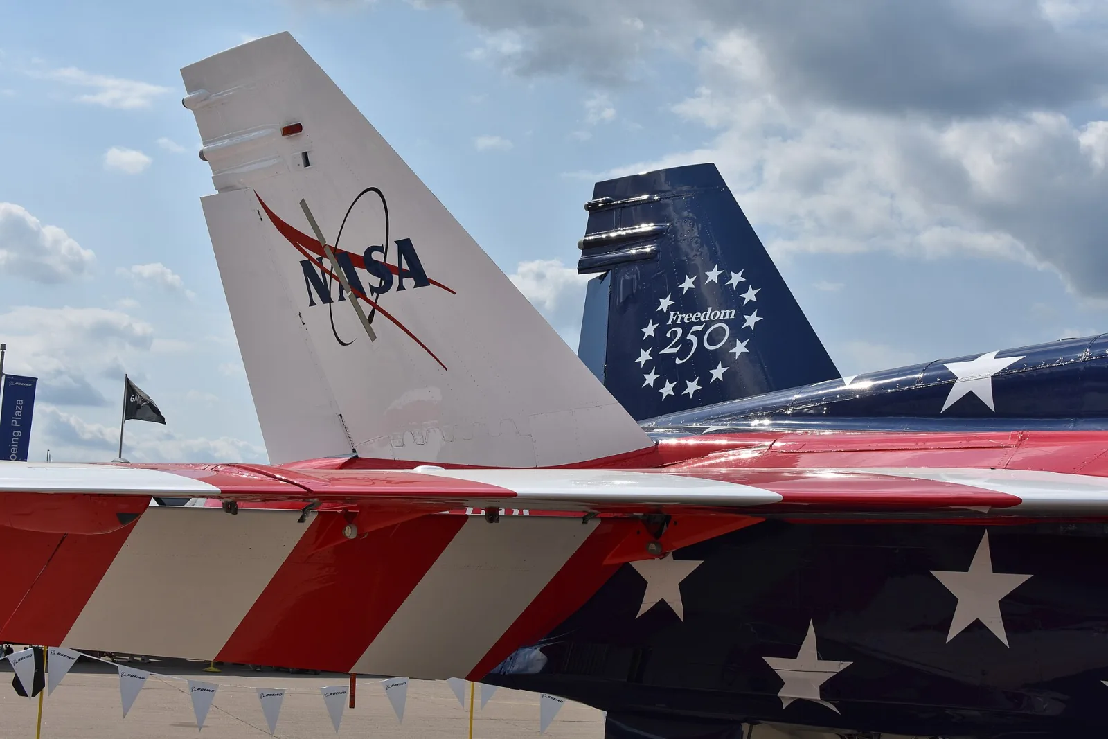 Deriva branca com o logotipo da NASA e asa com listras vermelhas e brancas de um F/A-18, com a deriva azul de outro jato com o emblema Freedom 250 ao fundo