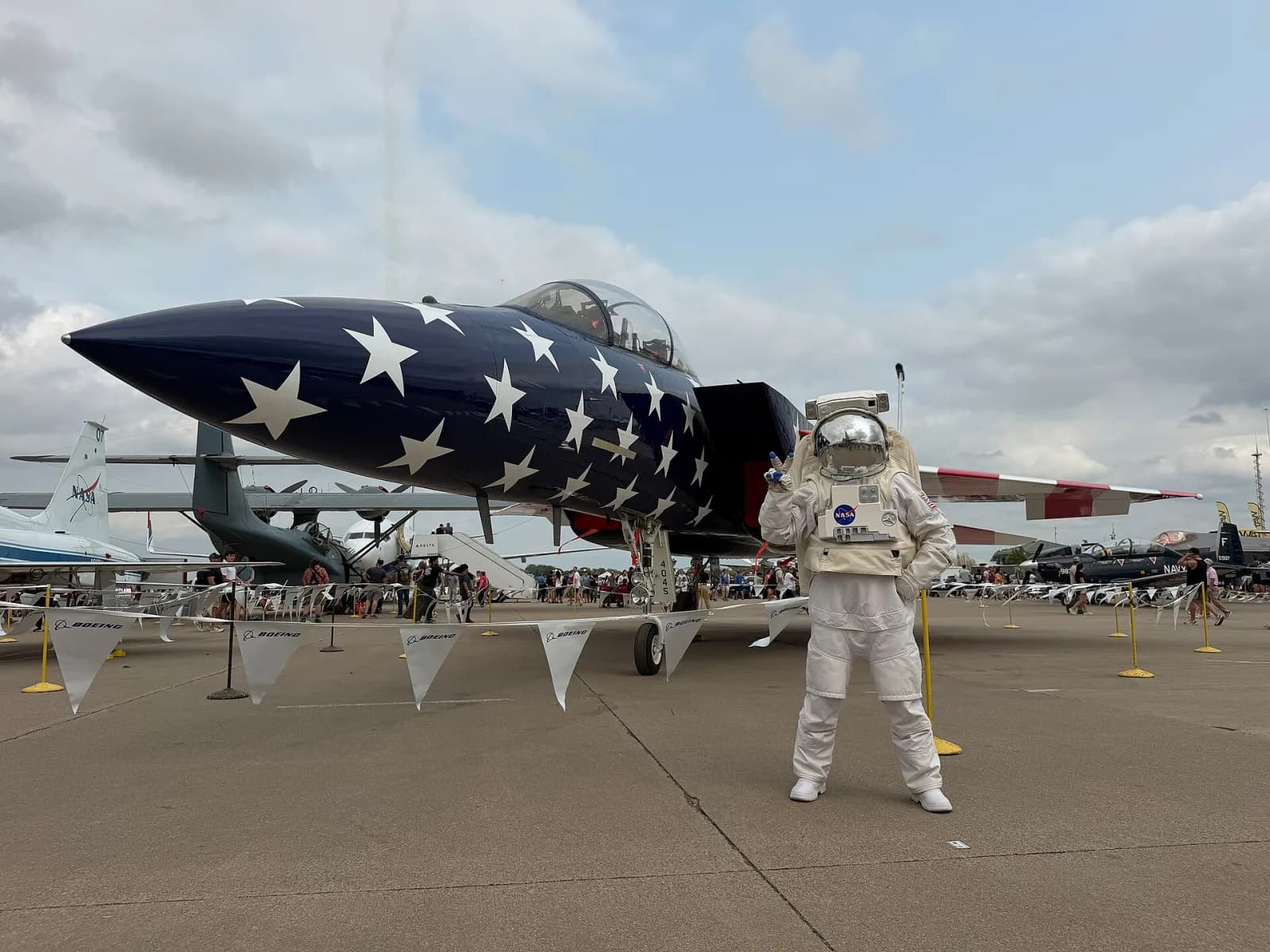 Caça F-15 com o nariz azul escuro coberto de estrelas brancas, estacionado no pátio com público ao fundo, e uma pessoa com traje de astronauta e viseira espelhada ao lado