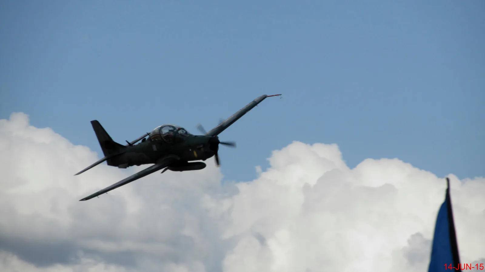 Avião militar de hélice com camuflagem verde em curva inclinada, contra nuvens brancas e céu azul