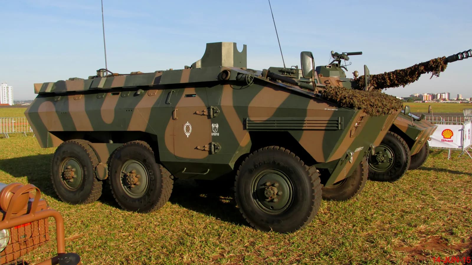 Dois blindados de rodas do Exército com camuflagem verde e marrom estacionados na grama, com grades de contenção ao fundo