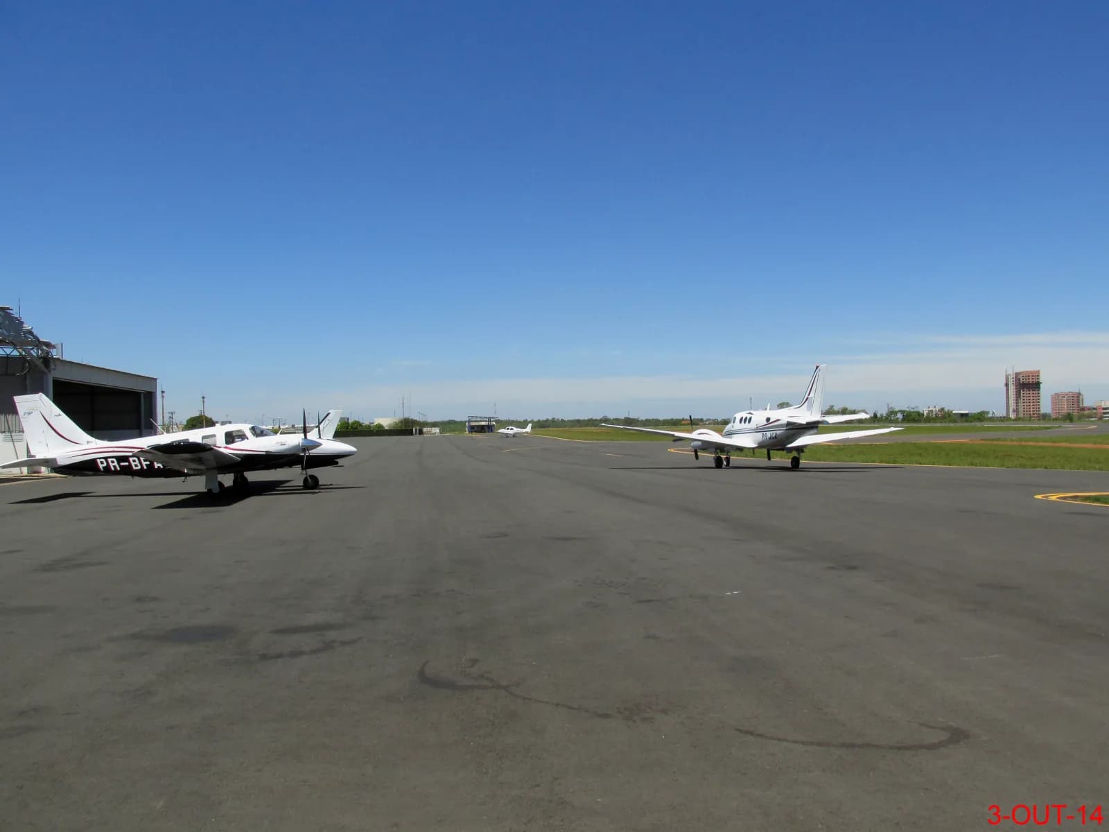 Pátio asfaltado de um aeródromo com dois aviões bimotores estacionados, um hangar à esquerda e céu azul limpo