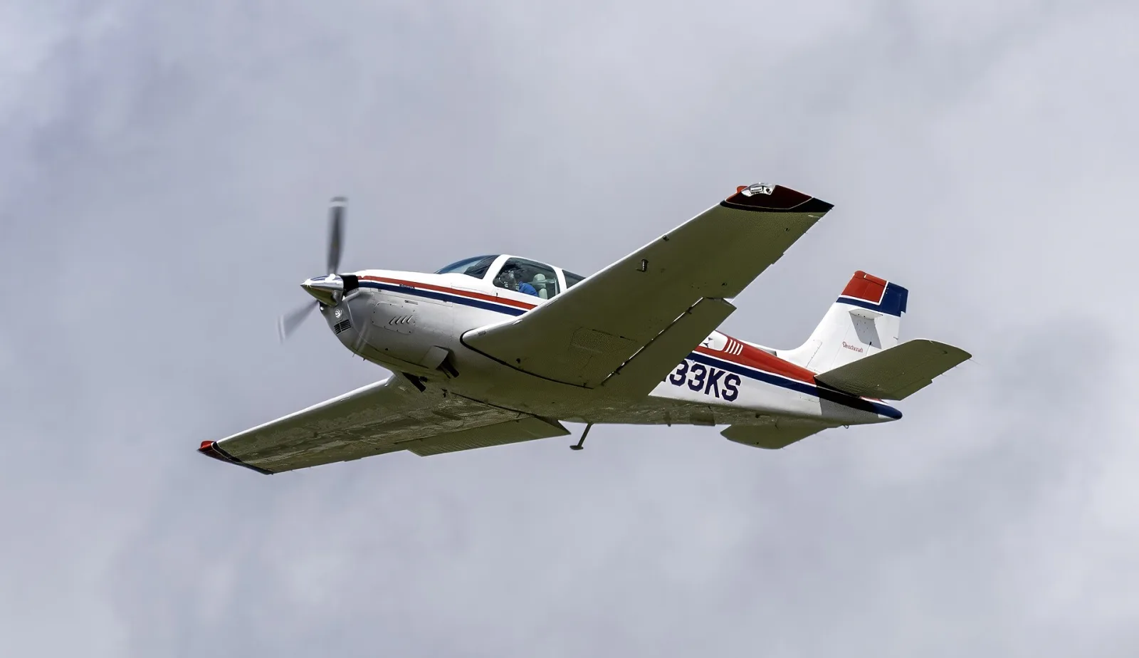 Beechcraft F33A Bonanza branco com faixas vermelha e azul em voo, de trem recolhido, contra céu nublado
