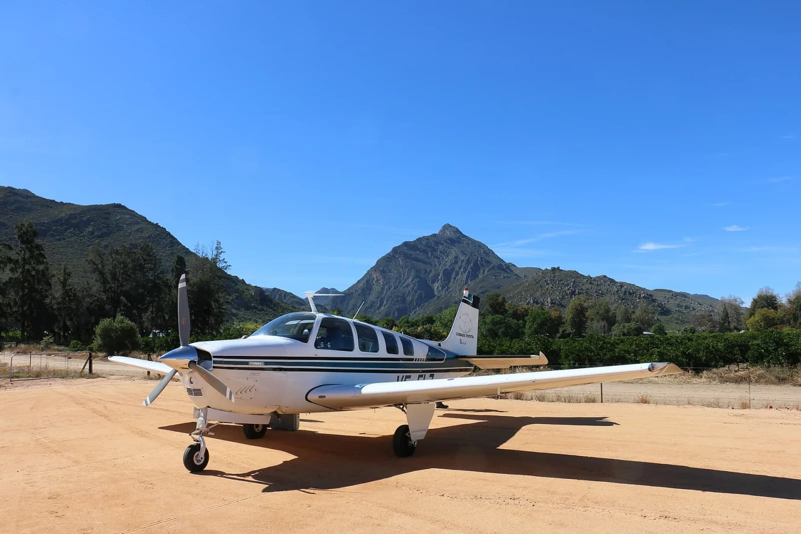 Beechcraft Bonanza parado numa pista de terra clara, com montanhas e céu azul ao fundo