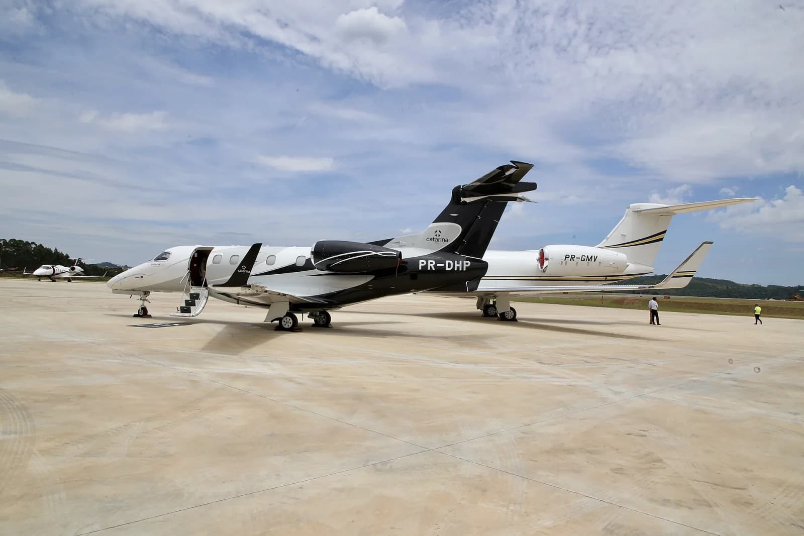 Jato Embraer Phenom 300 preto e branco com a marca Catarina na deriva, porta aberta, ao lado de um jato Gulfstream branco no pátio de concreto do aeroporto, com morros verdes ao fundo