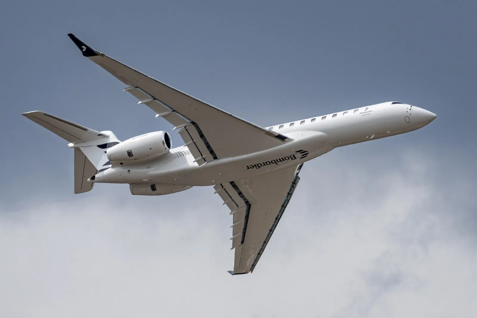Jato executivo Bombardier Global 8000 branco em voo, visto de baixo, com o nome Bombardier sob a fuselagem e céu nublado