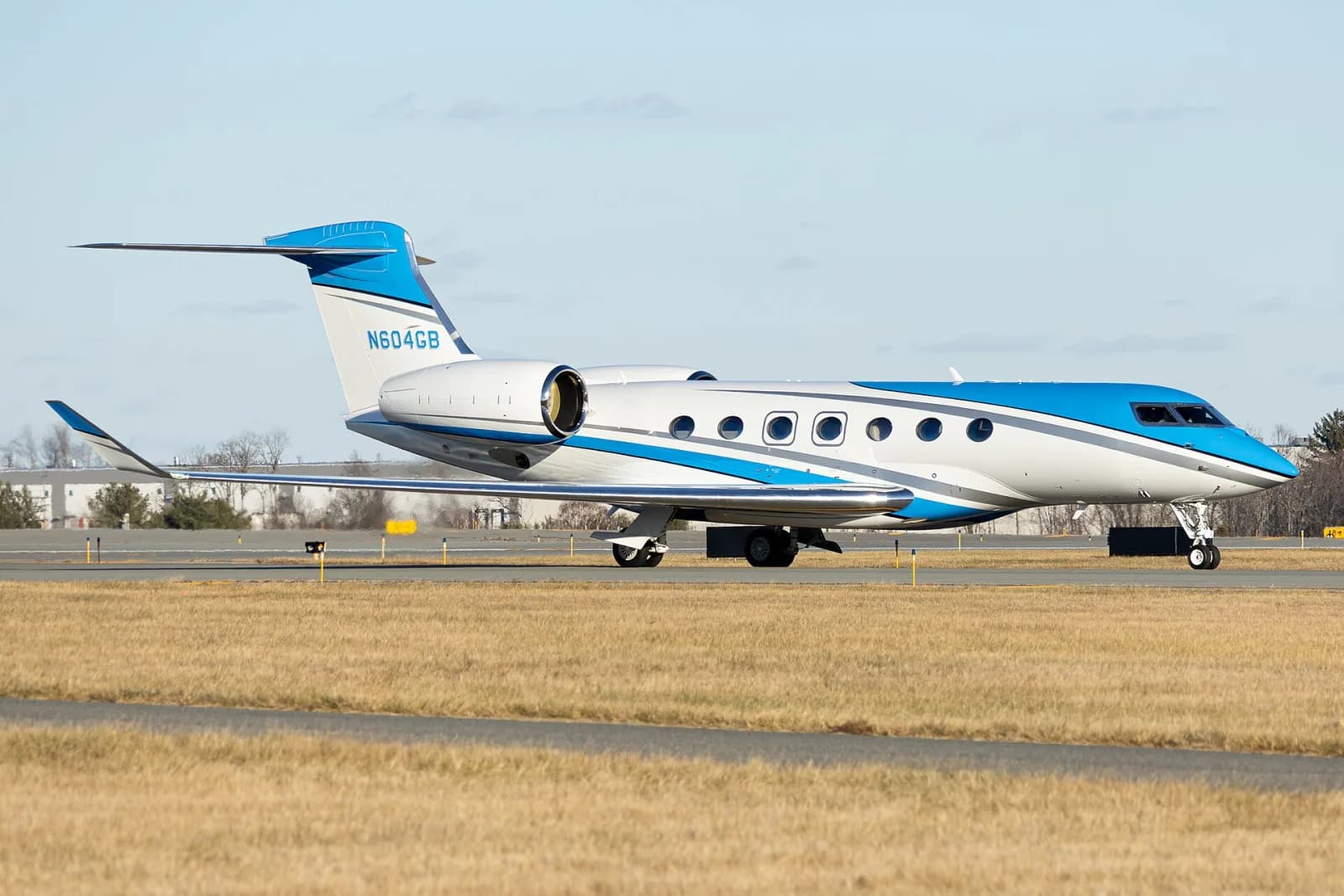 Jato executivo Gulfstream G600 branco com faixas azuis taxiando numa pista, com grama seca em primeiro plano