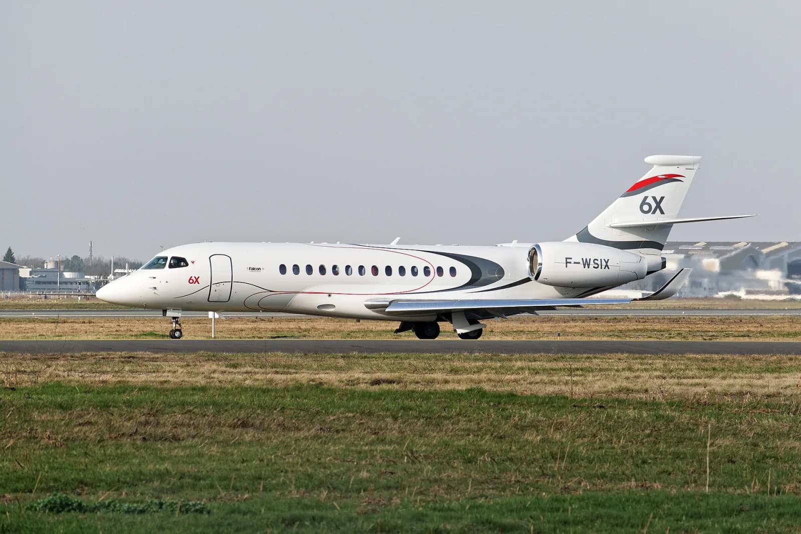 Jato executivo Dassault Falcon 6X branco, com «6X» pintado na deriva, taxiando numa pista com grama em primeiro plano