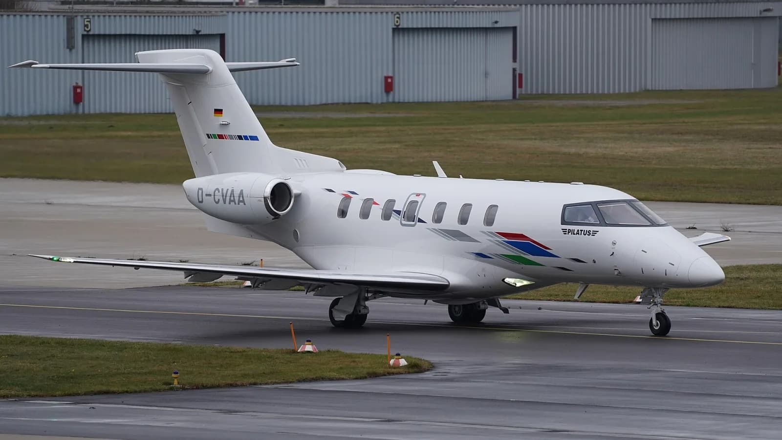 Jato executivo Pilatus PC-24 branco taxiando numa pista molhada, com hangares cinza ao fundo