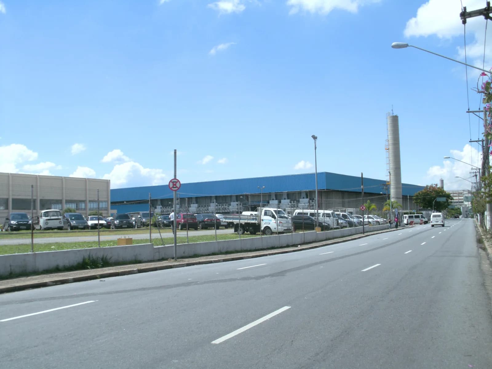 Avenida larga de quatro faixas com um pavilhão de telhado azul e um estacionamento de carros à esquerda, sob céu azul