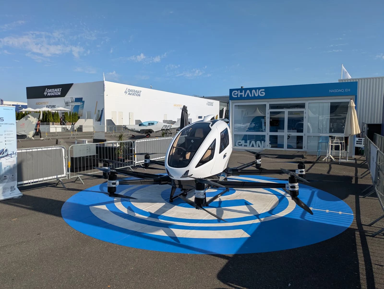 eVTOL EHang 216-S branco, de cabine fechada e oito braços com rotores, exposto sobre um círculo azul pintado no chão diante do estande da EHang