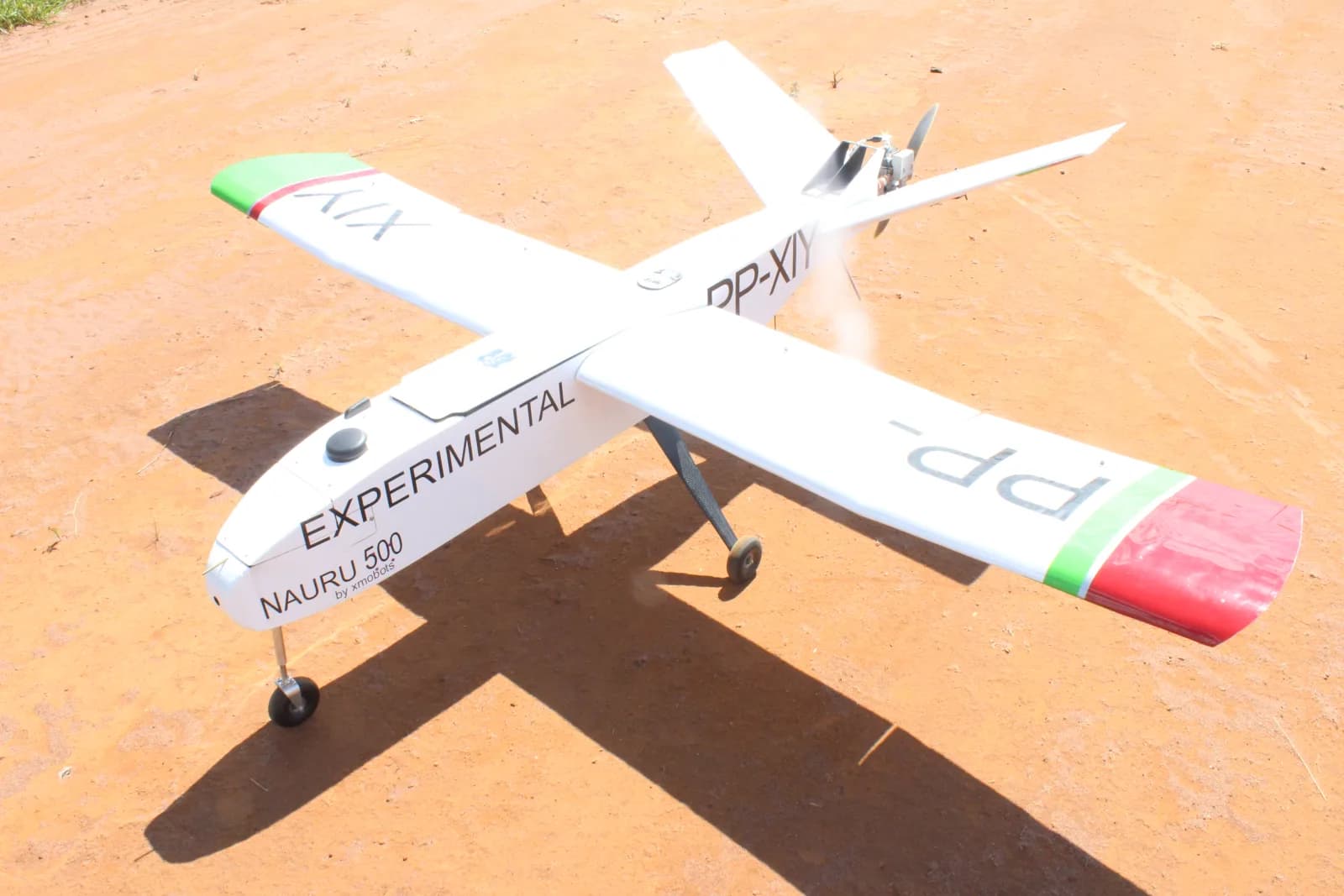 Drone de asa fixa branco Nauru 500, com a inscrição «Experimental» e pontas de asa verde e vermelha, parado sobre terra batida