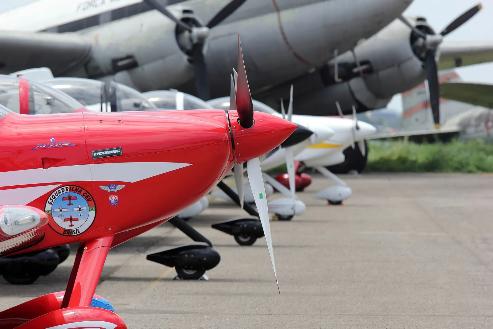 Nariz de um avião vermelho com o emblema da Esquadrilha CEU, com outros aviões da equipe e um avião antigo da FAB ao fundo