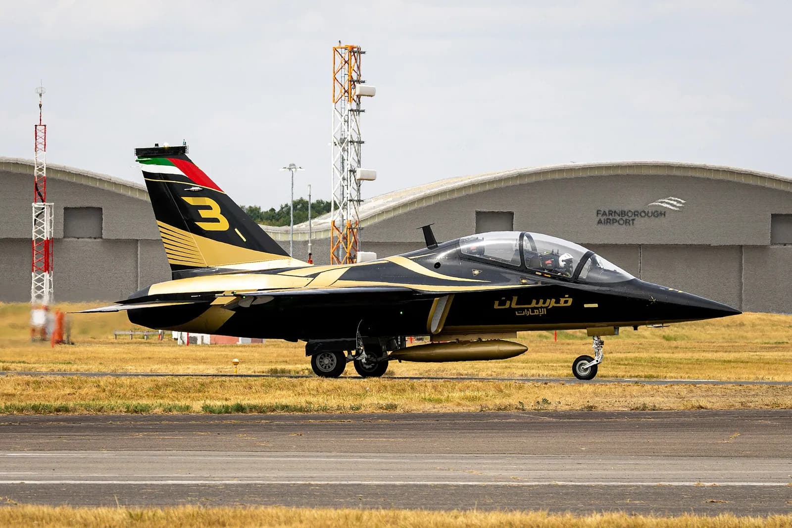 Jato preto e dourado com o número 3 na deriva taxiando diante de um hangar com a inscrição Farnborough Airport