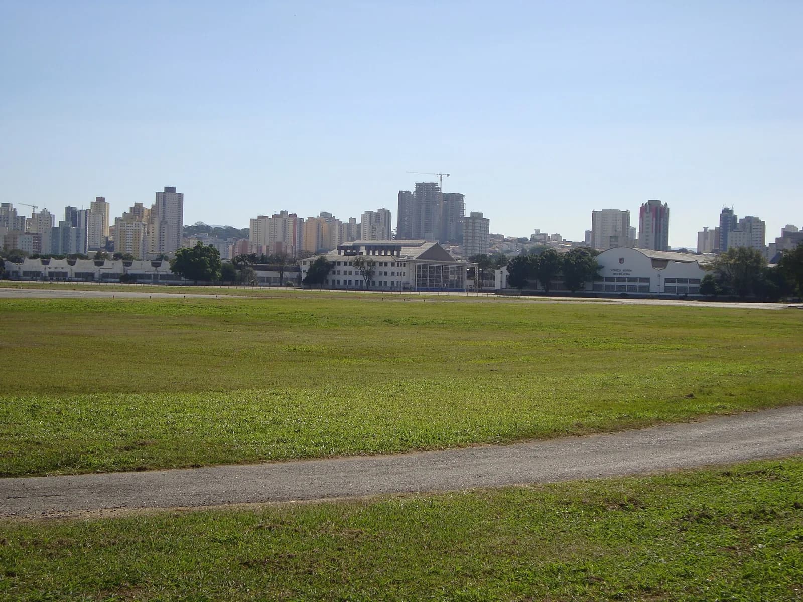 Gramado amplo do Campo de Marte com uma pista de taxiamento em primeiro plano, hangares brancos ao fundo, um deles com a inscrição «Força Aérea Brasileira», e prédios da zona norte de São Paulo no horizonte