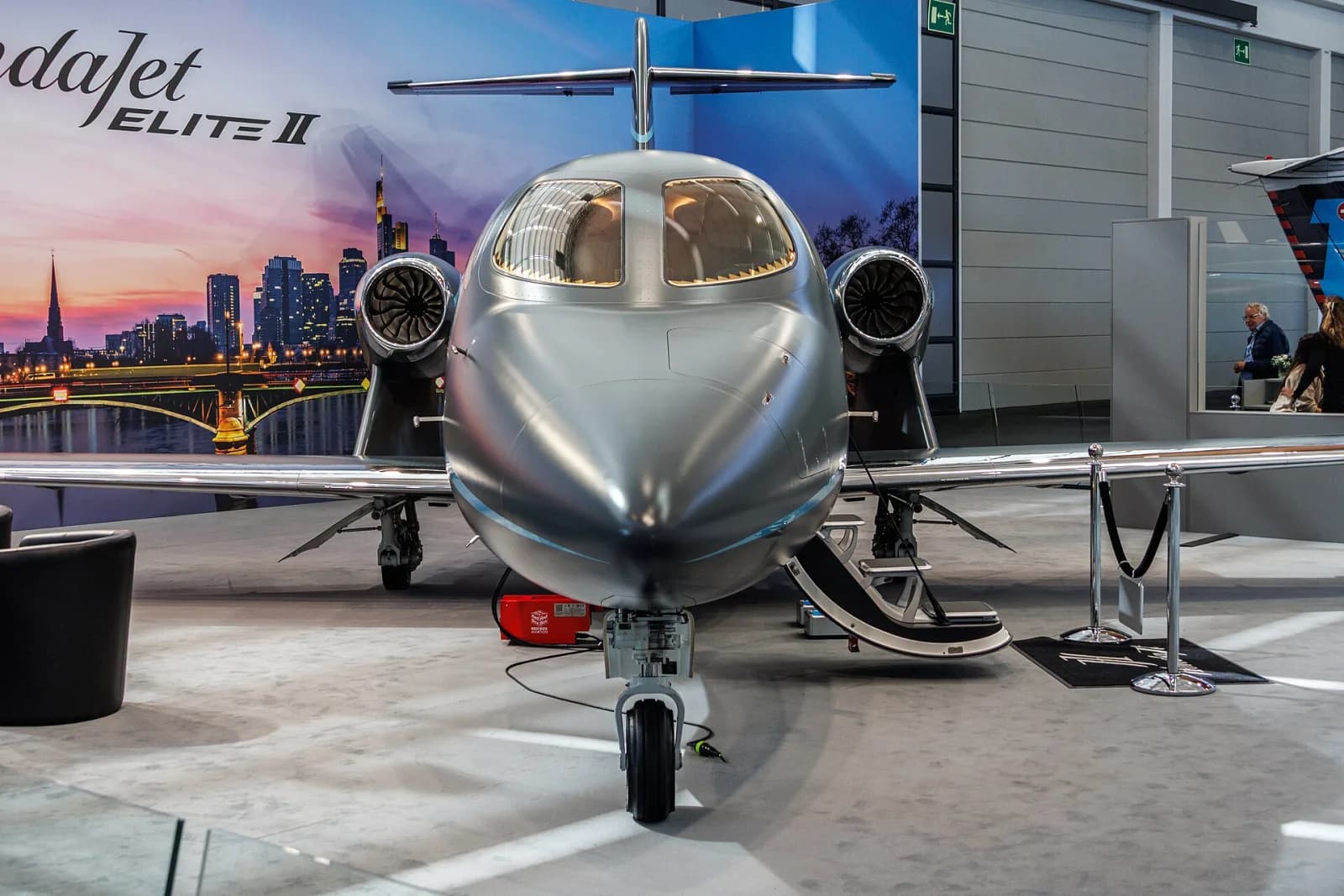 Jato executivo HondaJet Elite II prateado visto de frente, com a escada aberta, num estande de feira coberta com o painel «HondaJet Elite II» ao fundo