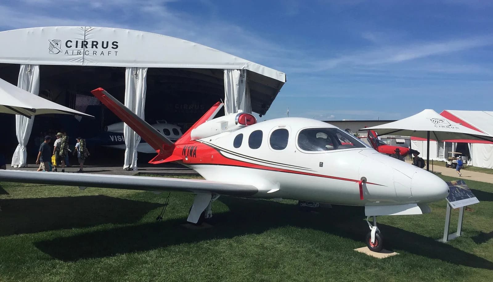 Jato monomotor Cirrus Vision SF50 branco e vermelho, com o motor sobre a fuselagem e a cauda em V, exposto na grama diante de uma tenda da Cirrus Aircraft