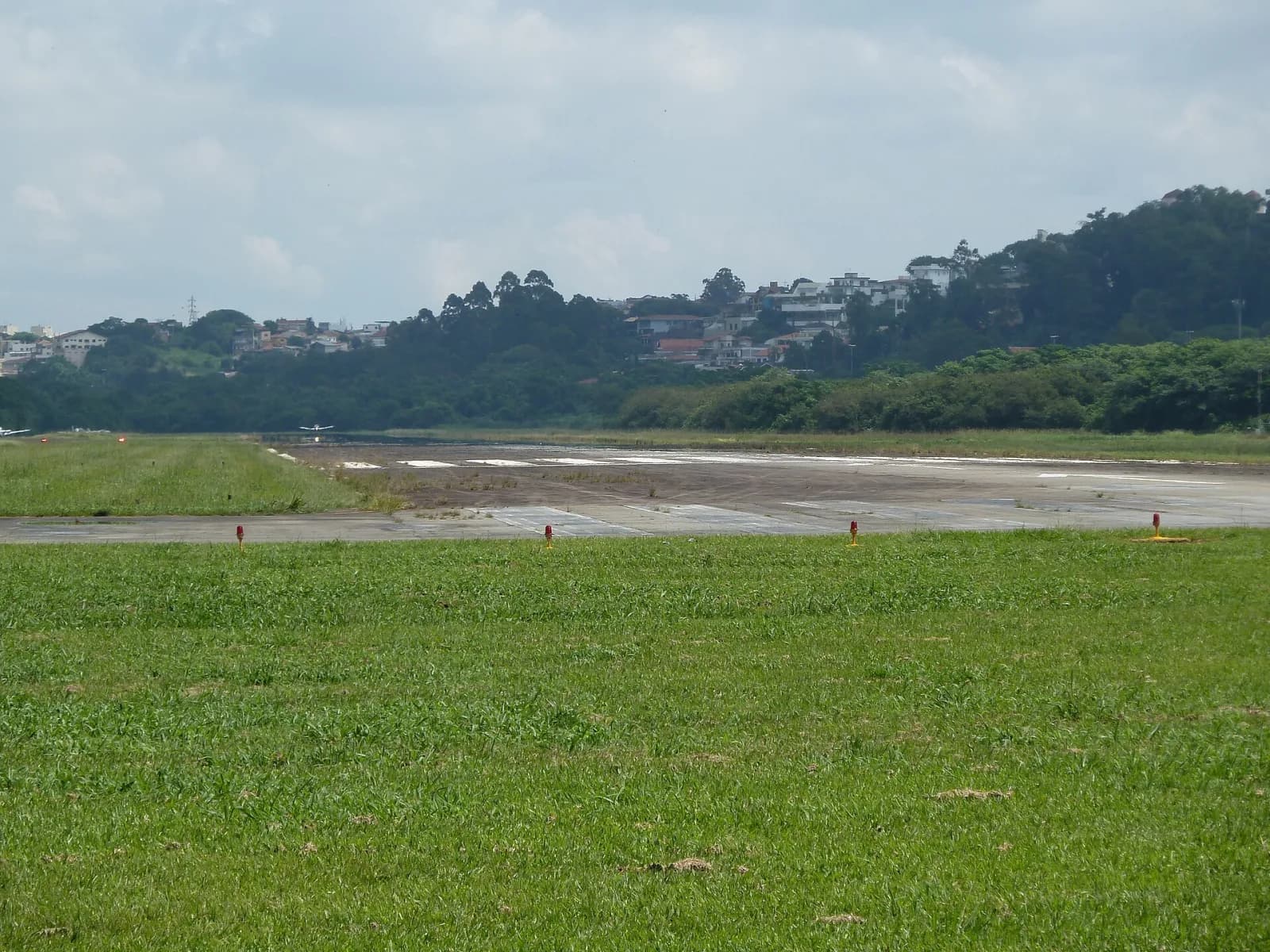 Pista de concreto do Campo de Marte vista da lateral, com gramado e luzes de balizamento em primeiro plano e morros com casas ao fundo