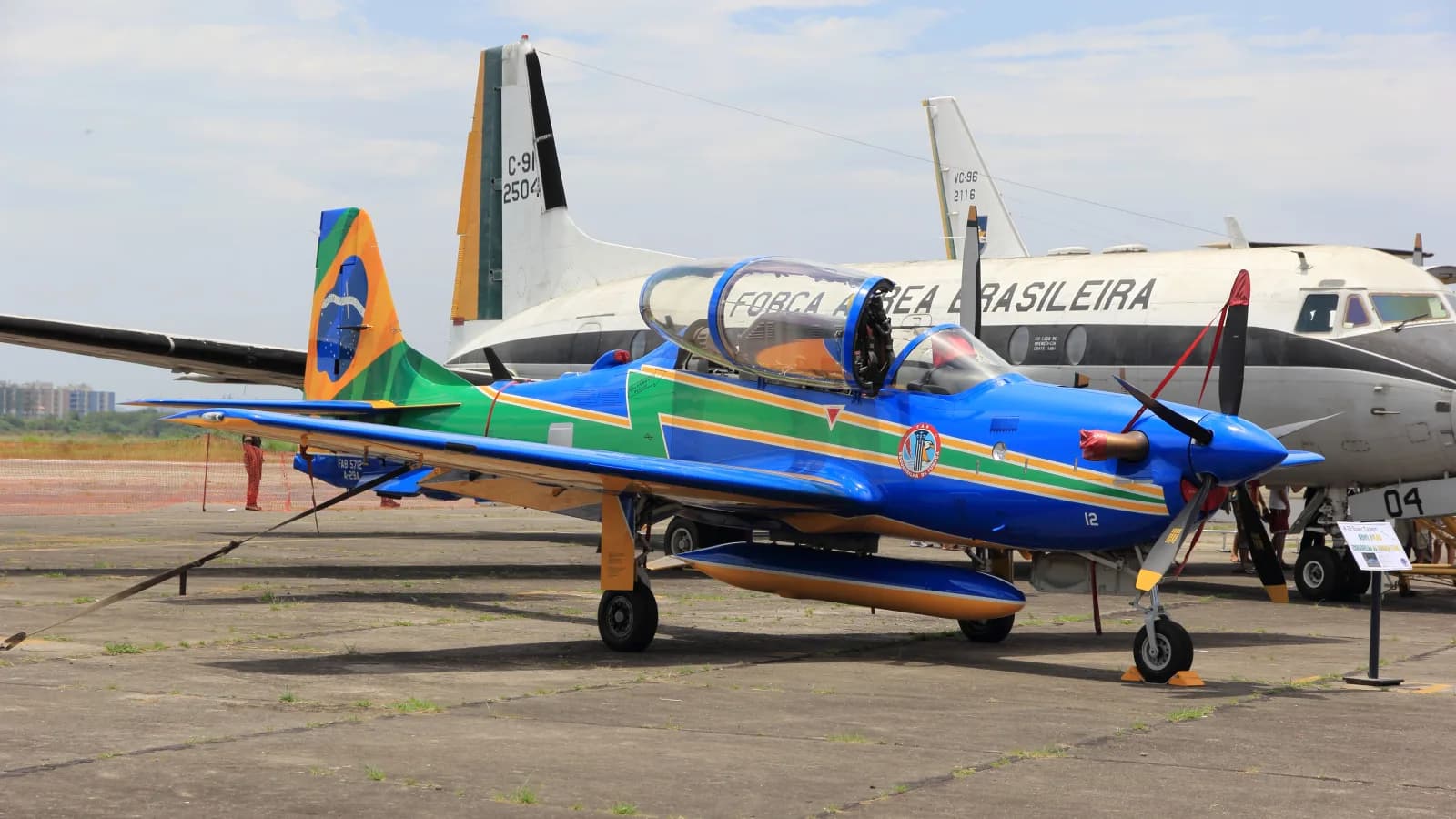 A-29 Super Tucano azul, verde e amarelo da Esquadrilha da Fumaça estacionado no pátio, com aviões de transporte históricos da FAB ao fundo