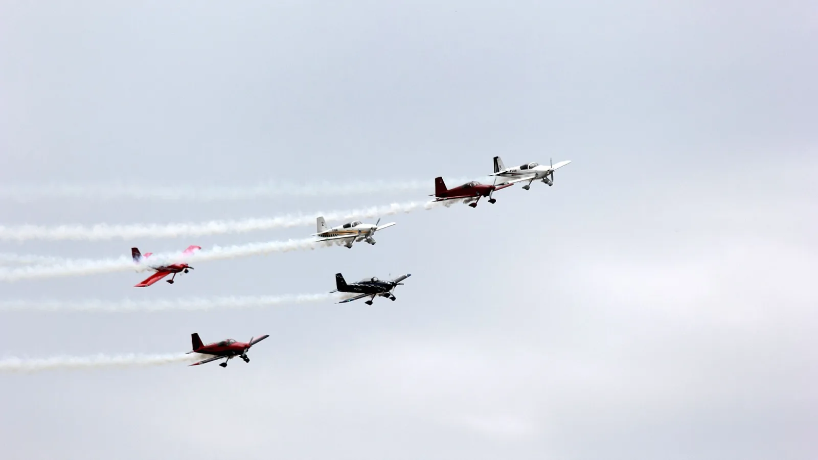 Seis aviões acrobáticos em formação deixando rastros de fumaça branca contra o céu nublado