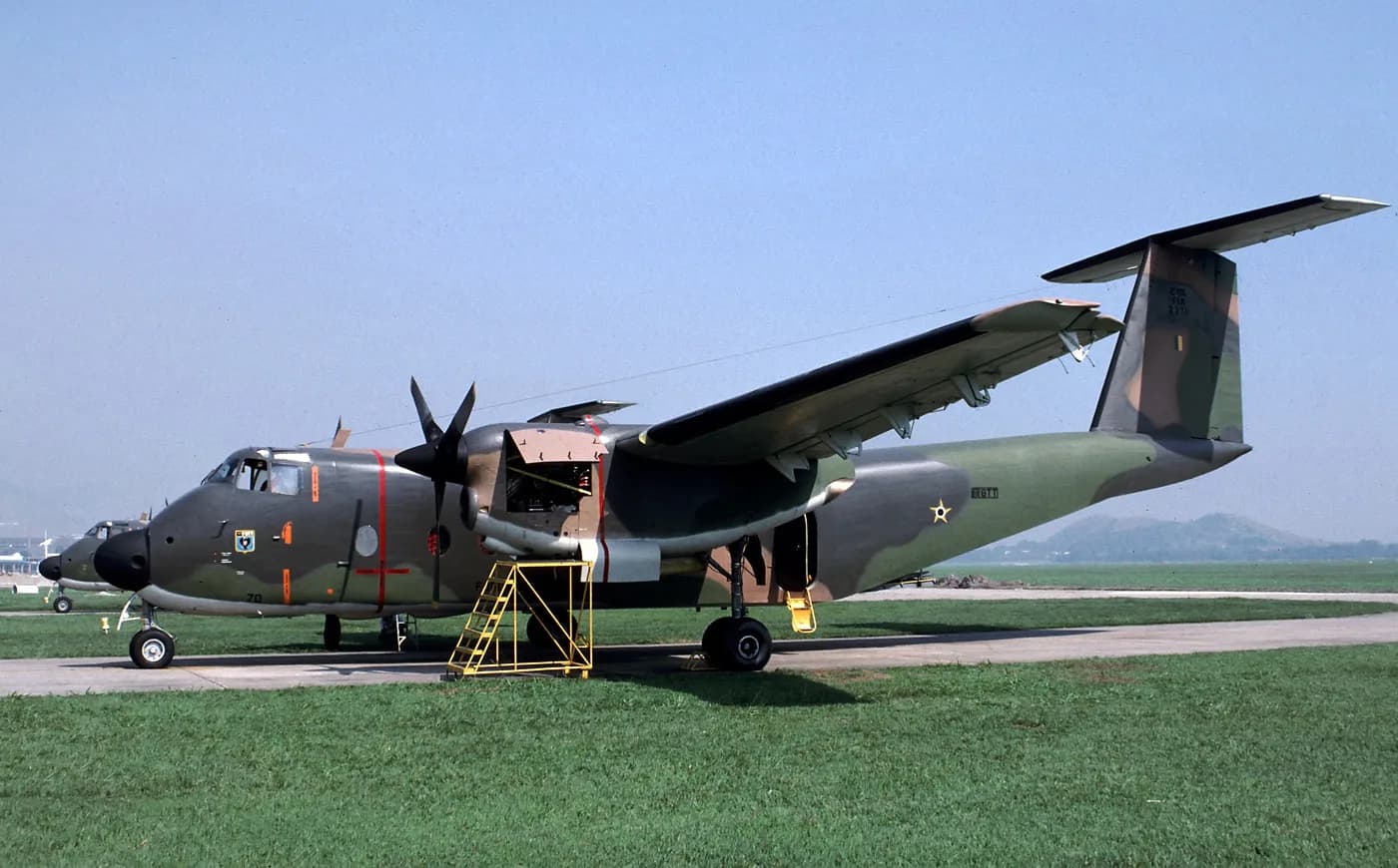 Avião de transporte C-115 Buffalo com camuflagem verde e marrom estacionado ao lado da grama de um aeródromo