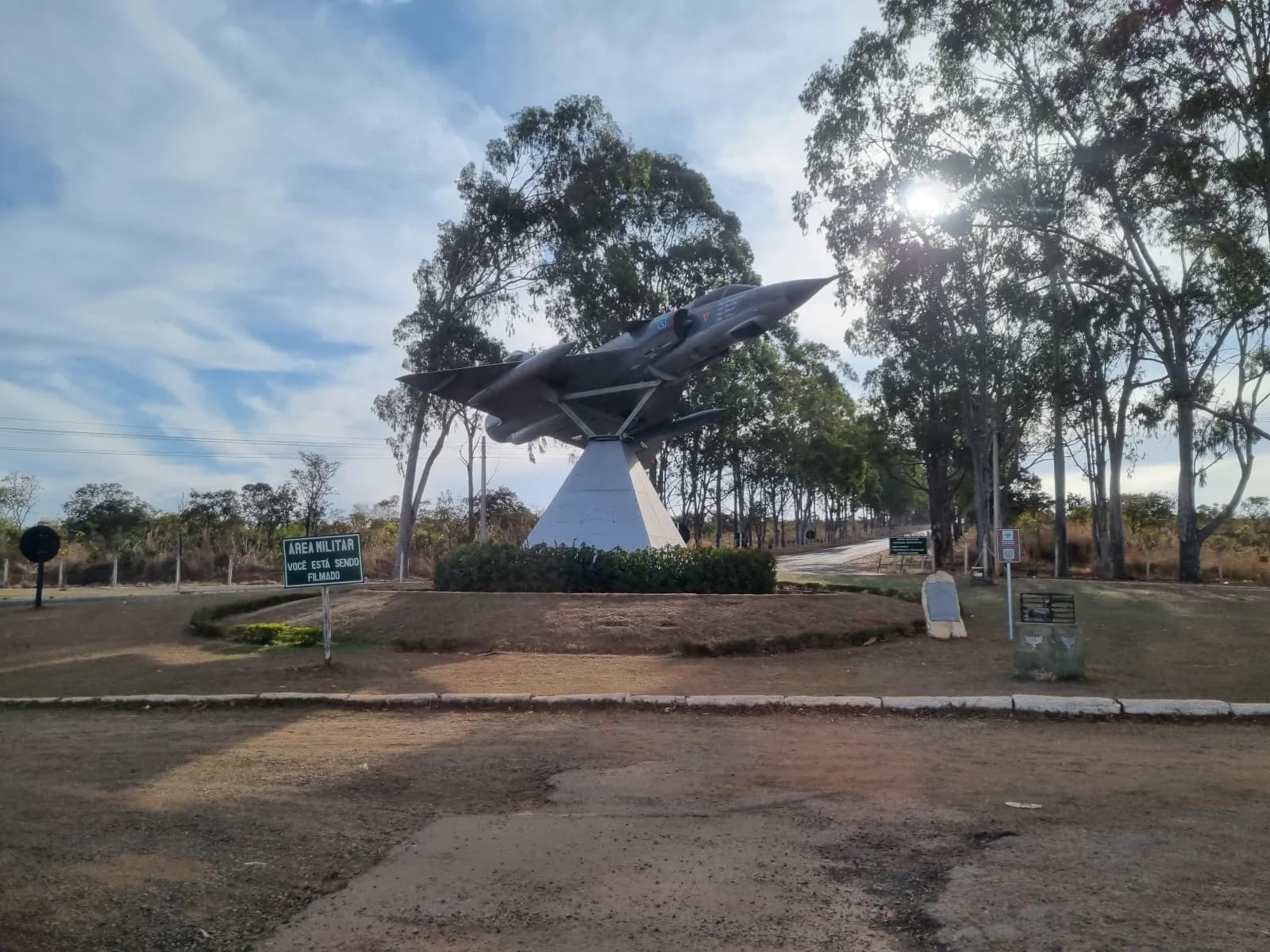 Caça Mirage montado sobre pedestal branco num canteiro gramado, com a placa «Área militar» à esquerda e eucaliptos ao fundo