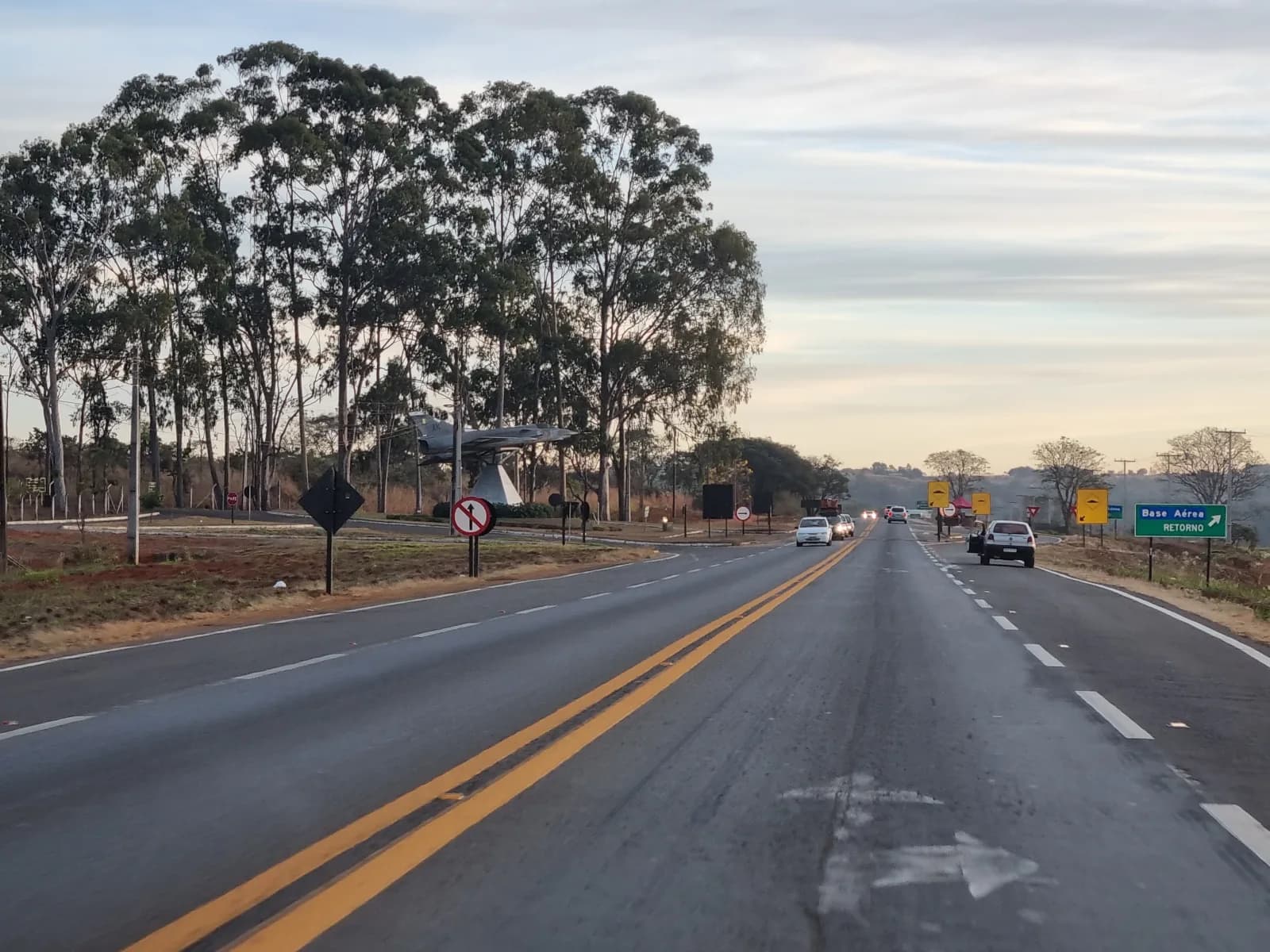 Rodovia de pista dupla com poucos carros, o monumento do Mirage entre eucaliptos à esquerda e a placa verde «Base Aérea, retorno» à direita