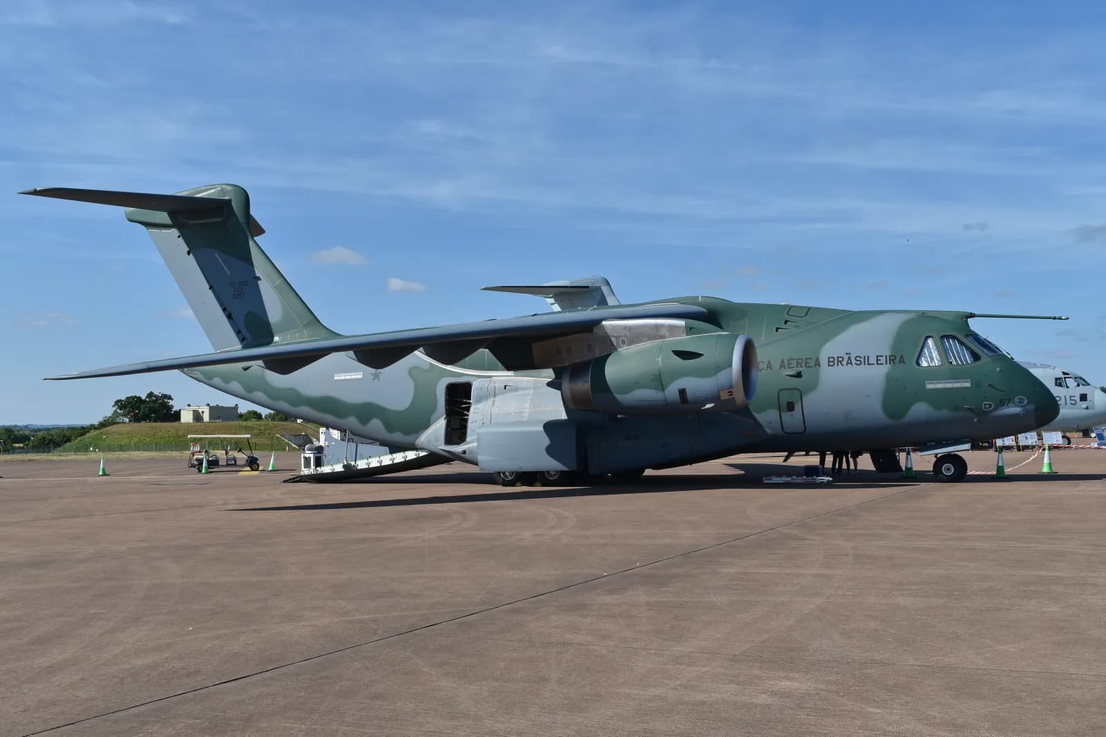 Cargueiro KC-390 Millennium da Força Aérea Brasileira, com pintura camuflada verde e cinza, estacionado num pátio com a porta lateral aberta