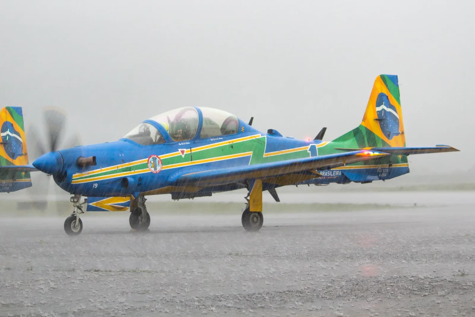 Avião A-29 Super Tucano da Esquadrilha da Fumaça, azul com faixas verde e amarela e a bandeira do Brasil na cauda, taxiando sob chuva forte