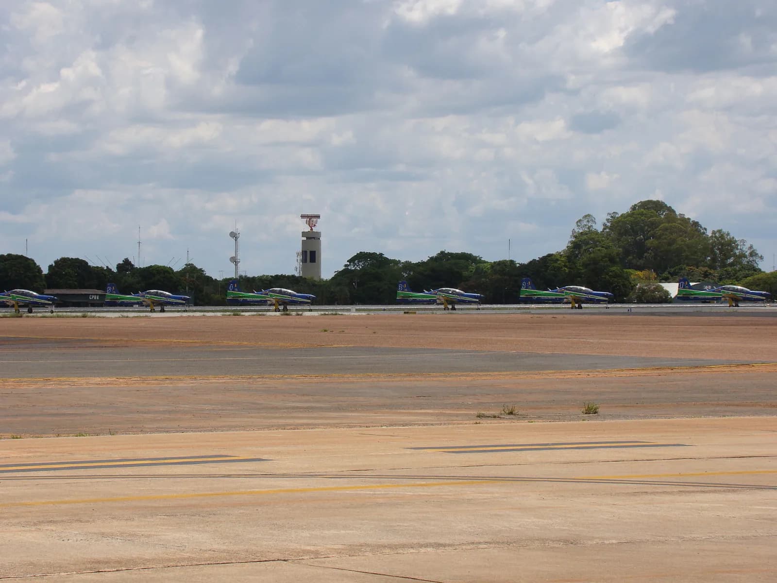 Fileira de aviões da Esquadrilha da Fumaça, azuis com faixas verde e amarela, estacionados num pátio largo, com uma torre e árvores ao fundo sob céu nublado