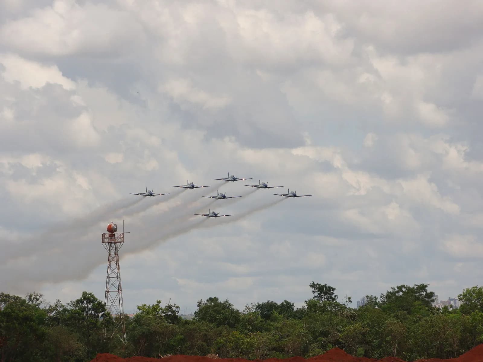Sete aviões da Esquadrilha da Fumaça em formação, soltando fumaça branca sobre o cerrado, com uma torre de sinalização em primeiro plano