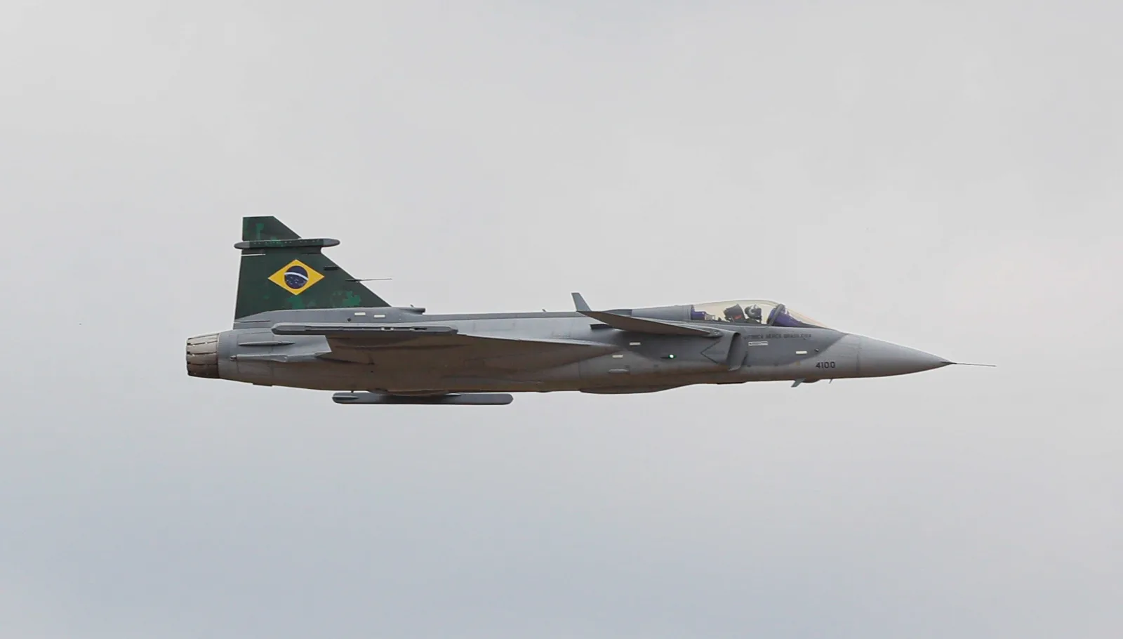 Caça F-39E Gripen com a bandeira do Brasil na deriva, em voo nivelado contra o céu cinza