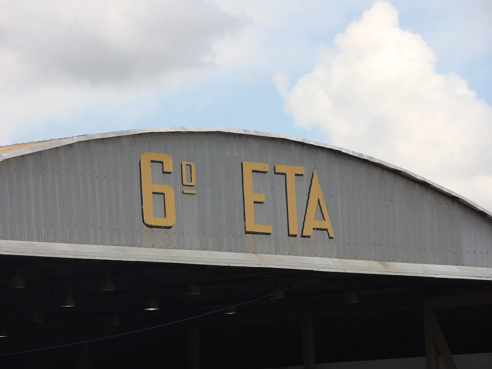 Fachada curva de um hangar metálico com a inscrição «6º ETA» em letras amarelas, sob céu azul com nuvens
