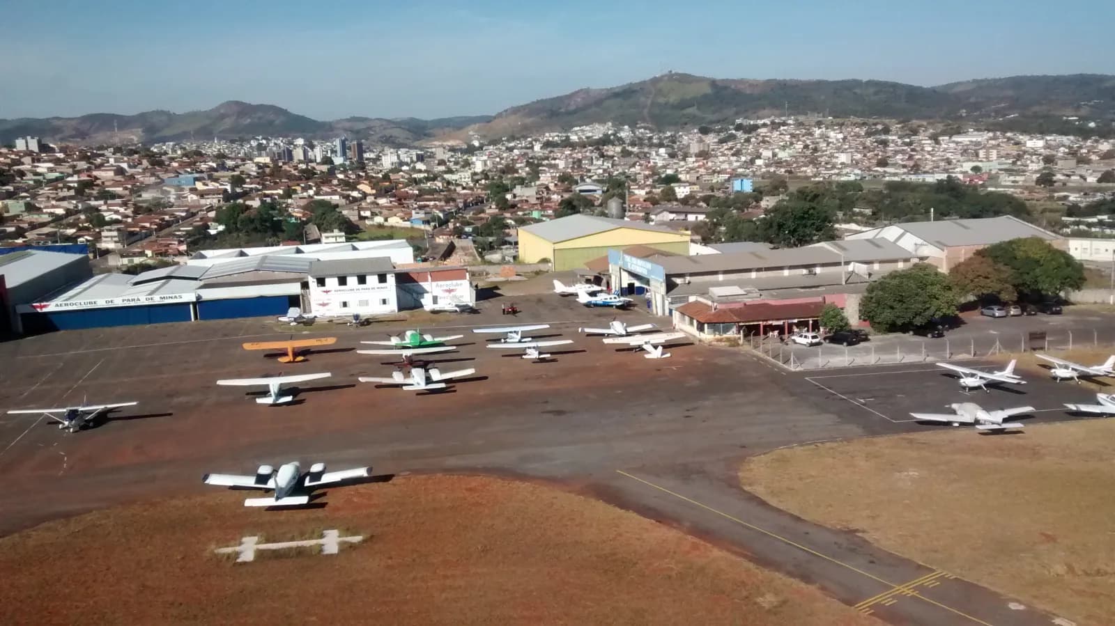 Vista aérea do pátio do Aeroclube de Pará de Minas, com aviões de pequeno porte estacionados, hangares e a cidade ao fundo