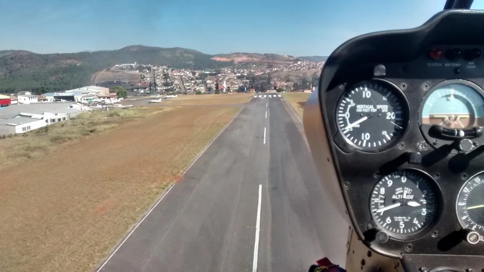 A pista asfaltada do aeródromo de Pará de Minas vista da cabine na aproximação, com o painel de instrumentos à direita e a cidade ao fundo