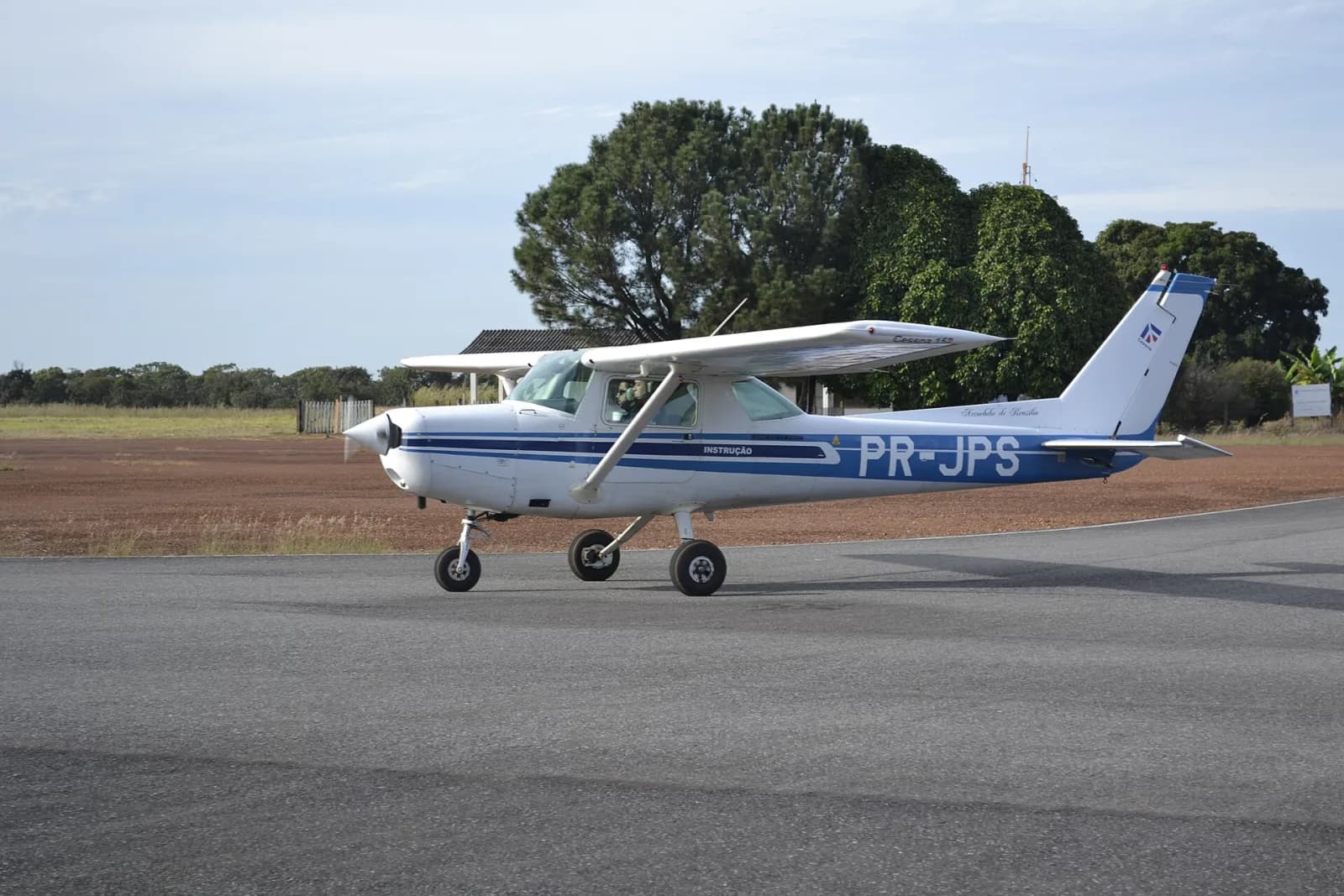 Cessna 152 branco e azul de matrícula PR-JPS, com a inscrição Instrução, taxiando numa pista com árvores ao fundo