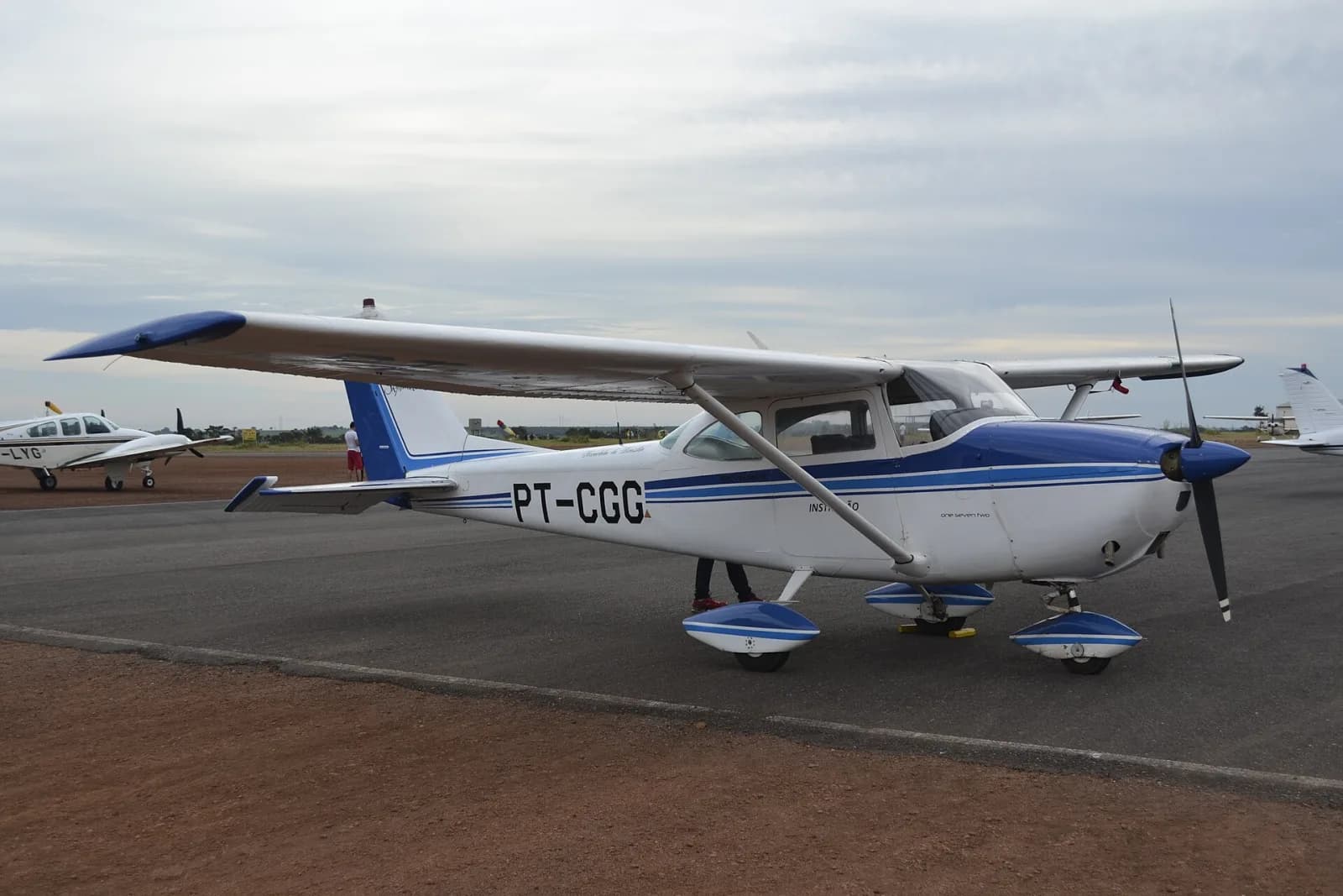 Cessna 172 branco e azul de matrícula PT-CGG estacionado no pátio, sob céu nublado, com outros aviões ao fundo
