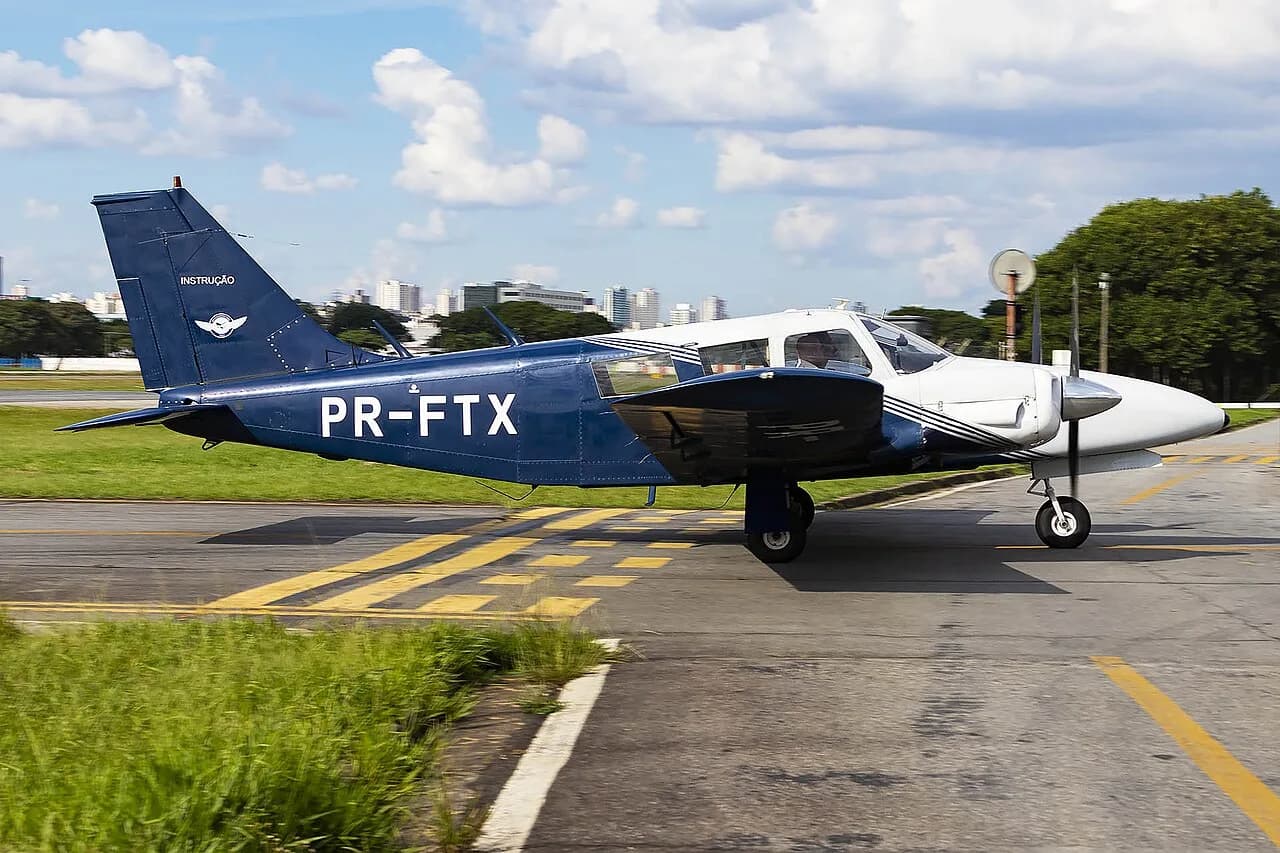 Bimotor Piper Seneca azul e branco de matrícula PR-FTX taxiando, com prédios e árvores ao fundo