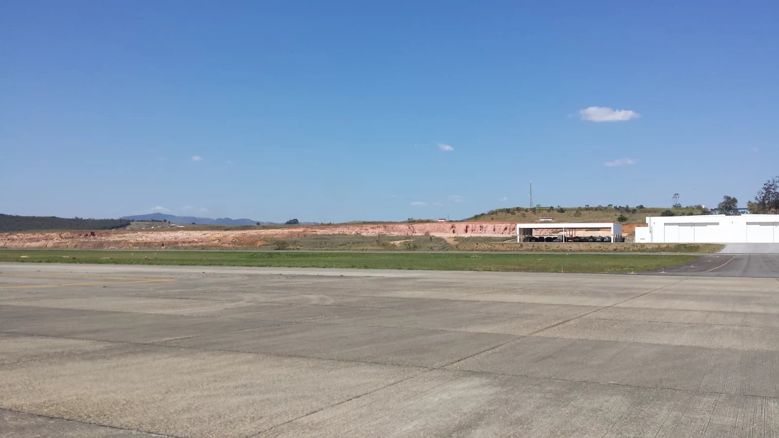 Pátio de concreto e pista da Base de Aviação de Taubaté, com hangares brancos à direita e morros do Vale do Paraíba ao fundo