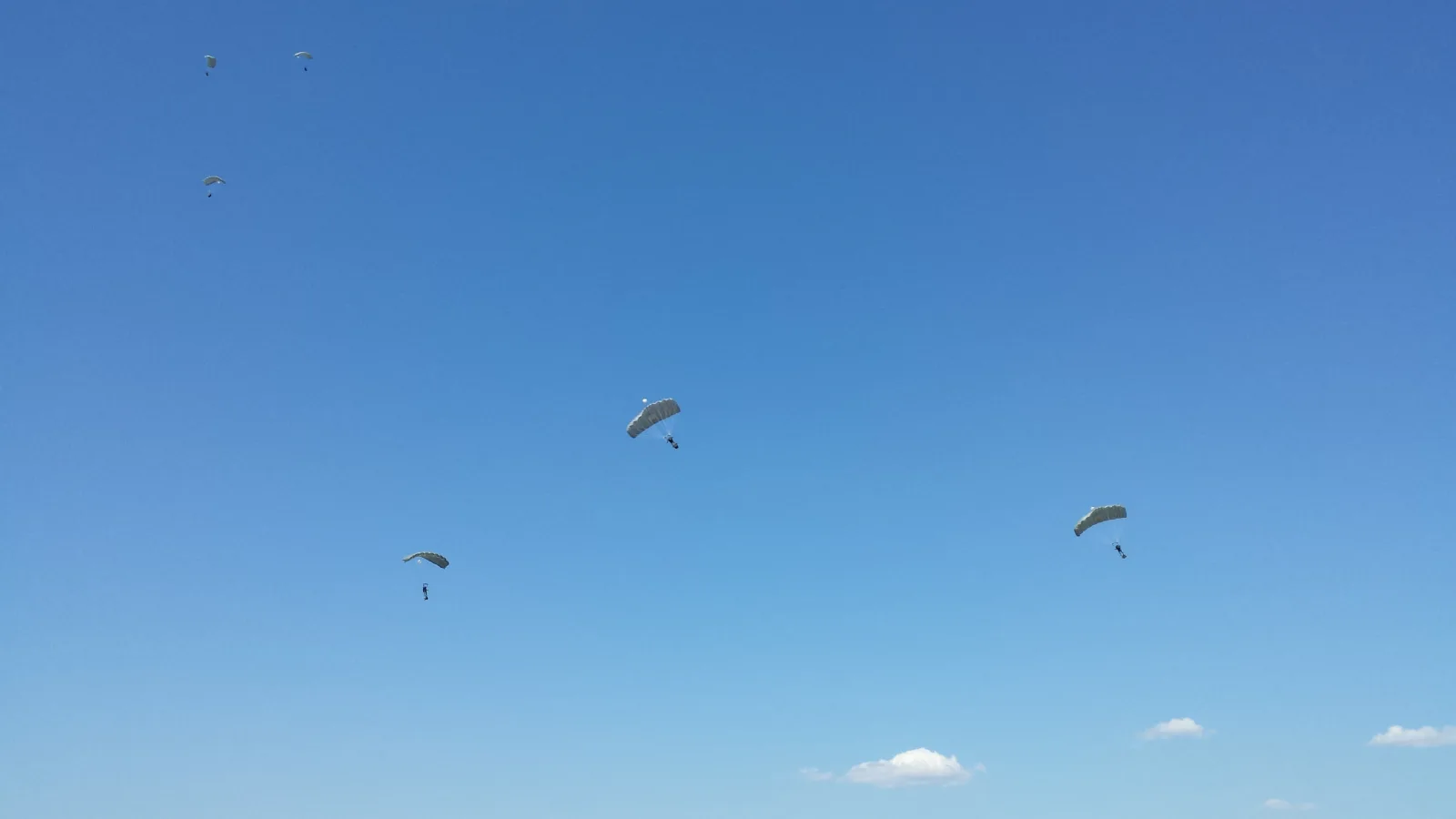 Seis paraquedistas militares com velames verde-claros descendo contra o céu azul