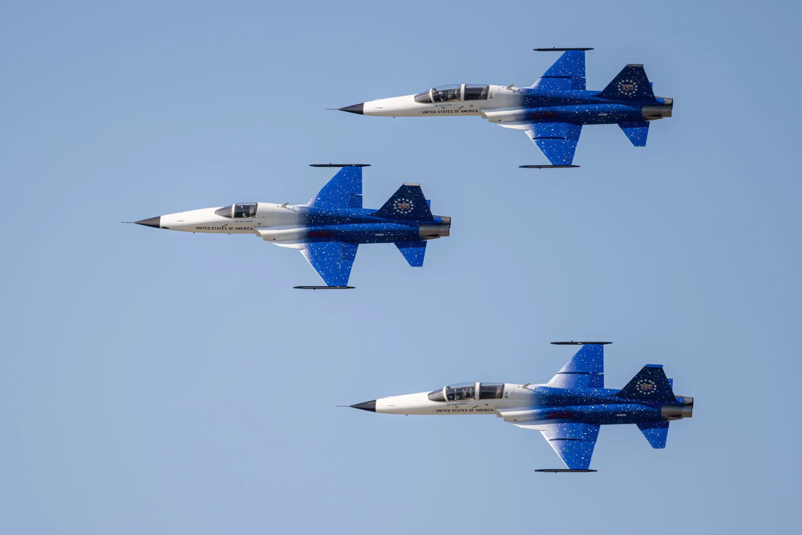 Três caças F-5 brancos e azuis, com a inscrição United States of America e o emblema dos 250 anos na deriva, voando em formação contra o céu azul