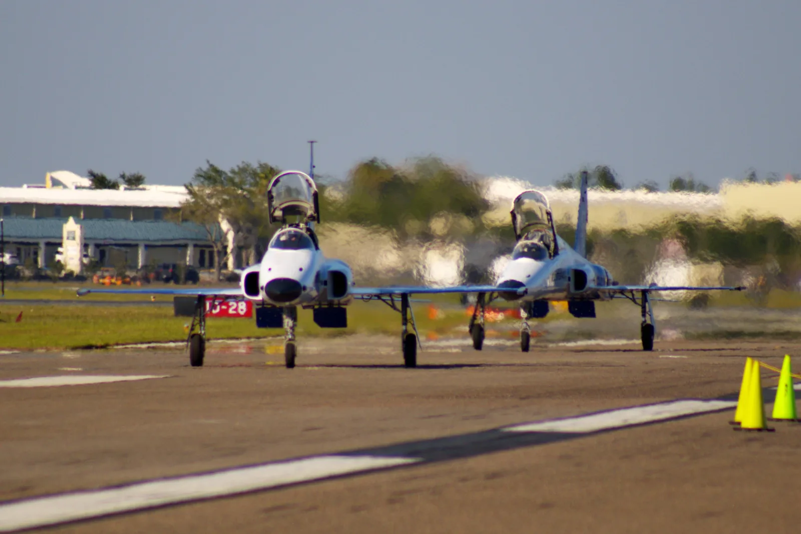 Dois caças F-5 vistos de frente, com as capotas abertas, taxiando lado a lado na pista do aeroporto de Lakeland, com cones amarelos no canto