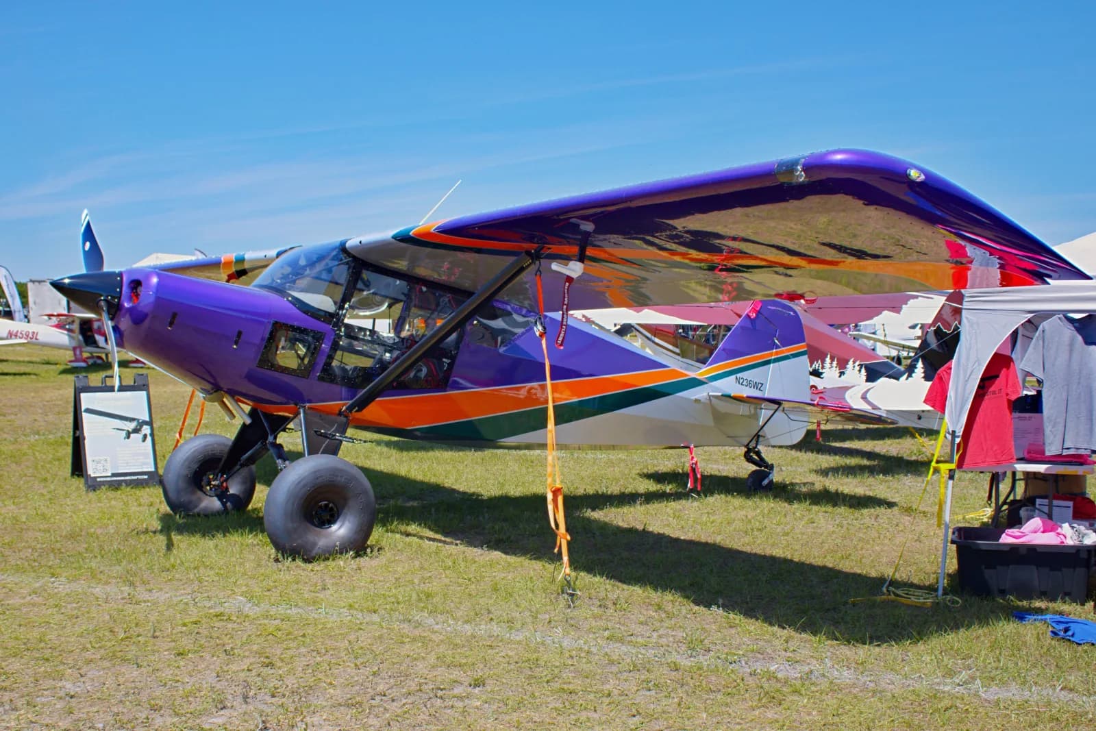 Monomotor de asa alta Bearhawk Companion roxo, com pneus grandes, estacionado na grama da área de exposição, com outros aviões e uma barraca ao fundo