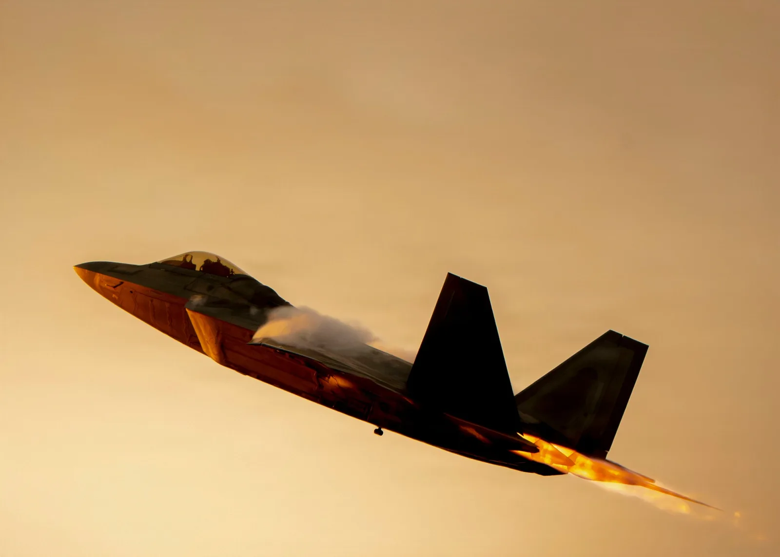 Caça F-22 Raptor subindo com o pós-combustor aceso e vapor sobre as asas, contra o céu alaranjado do fim de tarde