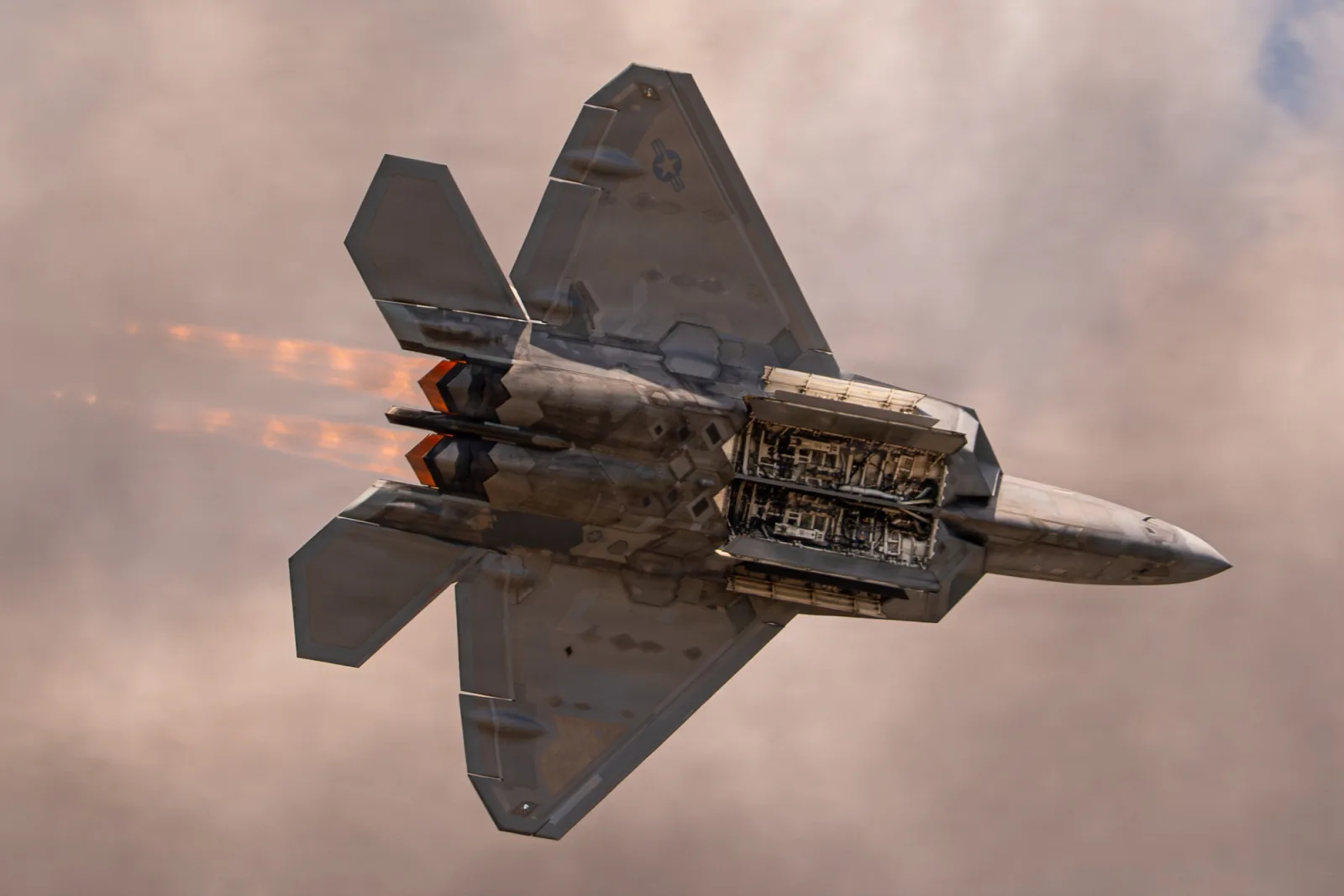 F-22 Raptor visto por baixo, com as portas do compartimento de armas abertas e o pós-combustor aceso, contra nuvens rosadas