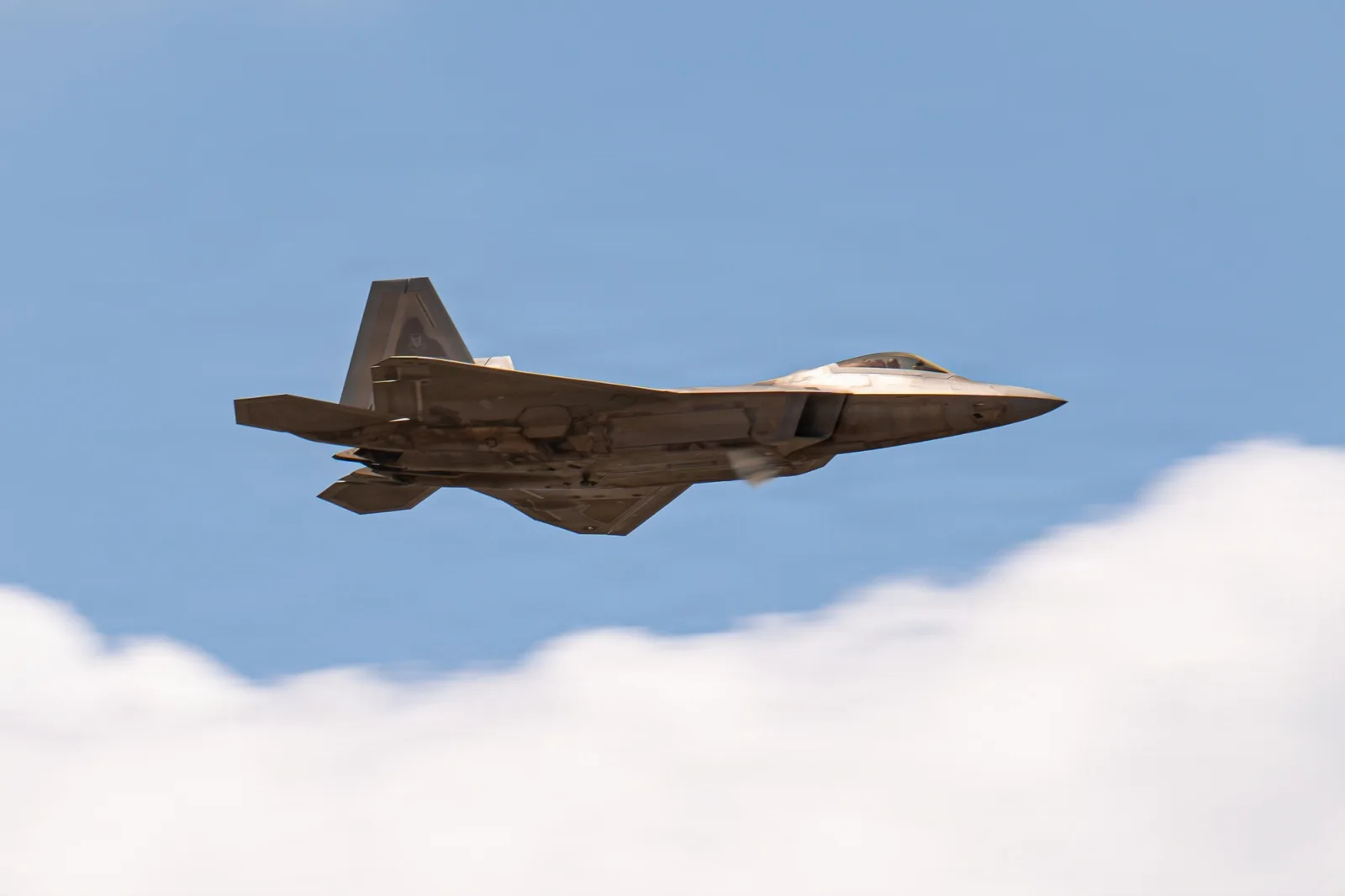 F-22 Raptor de perfil, em voo nivelado contra o céu azul, com uma nuvem branca embaixo